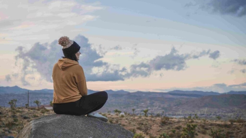 woman wearing brown hoodie. black leggins and a christmas beanie sitting on a rock looking over to the panoramic view